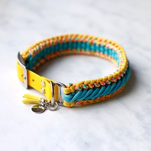 May include: A pet collar featuring a yellow strap with a silver buckle and D-ring. The collar is decorated with braided paracord in yellow, teal, and a multi-colored pattern. A small yellow tassel and a round silver metal tag are attached.