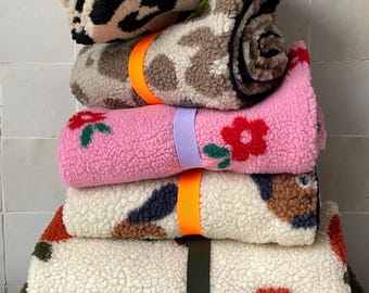 May include: A stack of six rolled sherpa fleece blankets featuring various patterns including leopard print, pink with red flowers, and abstract shapes in orange, green, and brown. Each blanket is secured with a colorful elastic strap.
