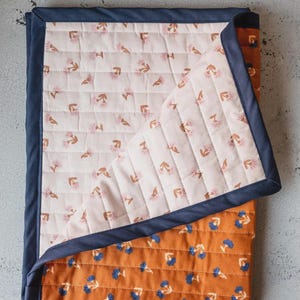 May include: A quilted blanket featuring a reversible design with a dark navy blue fabric border. One side displays a light cream background with small pink flower motifs, while the other side shows a burnt orange background with blue flowers.