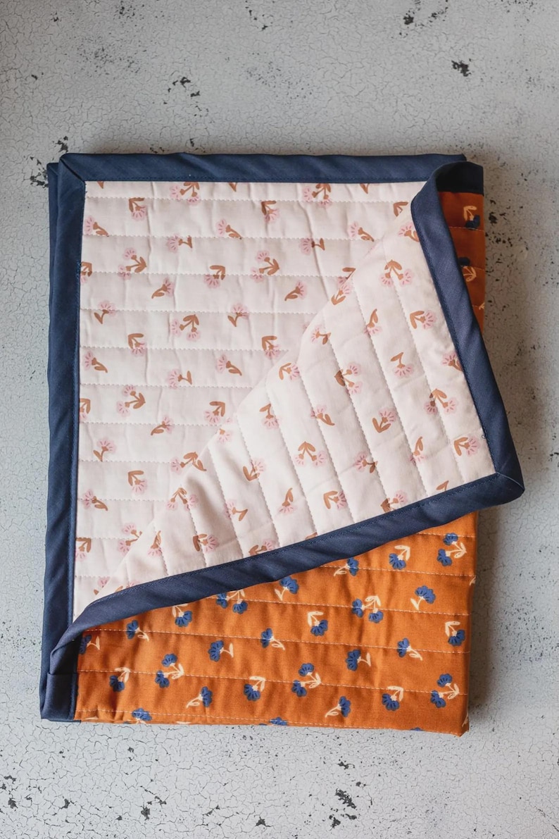 May include: A quilted blanket featuring a reversible design with a dark navy blue fabric border. One side displays a light cream background with small pink flower motifs, while the other side shows a burnt orange background with blue flowers.
