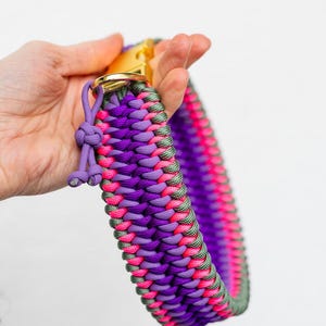 May include: A vibrant dog collar in shades of purple, pink, and green, with a gold-colored metal clasp. The collar is made of woven paracord, showcasing a detailed braided pattern. A small, knotted detail adds a decorative touch.