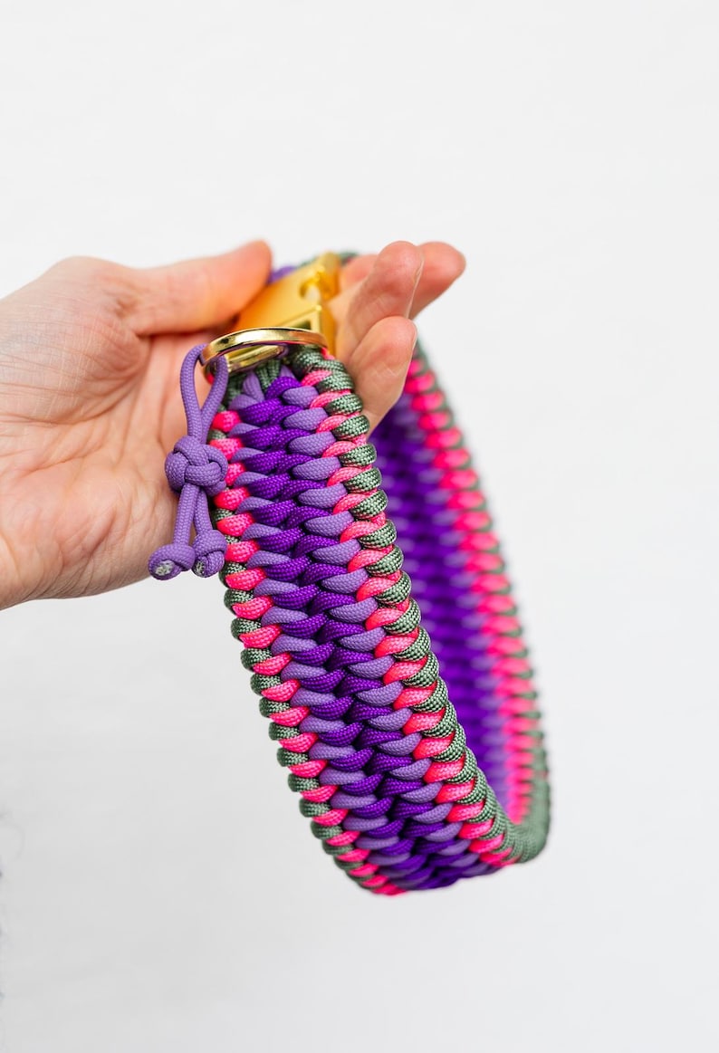 May include: A vibrant dog collar in shades of purple, pink, and green, with a gold-colored metal clasp. The collar is made of woven paracord, showcasing a detailed braided pattern. A small, knotted detail adds a decorative touch.