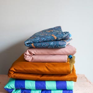 May include: A stack of folded fabric pieces. The top layer is dark blue with a fox and floral print. Below it is a solid dusty pink piece, followed by a solid mustard yellow piece, and a bottom layer featuring bold teal and purple stripes.