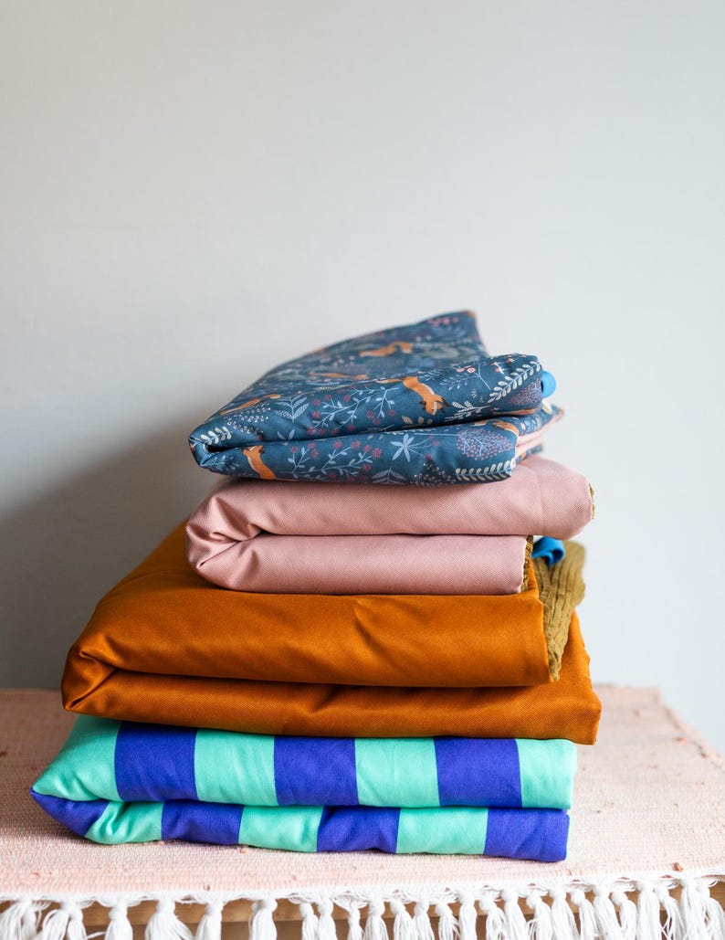 May include: A stack of folded fabric pieces. The top layer is dark blue with a fox and floral print. Below it is a solid dusty pink piece, followed by a solid mustard yellow piece, and a bottom layer featuring bold teal and purple stripes.
