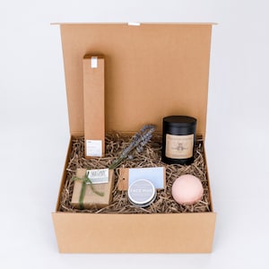 May include: A brown cardboard gift box filled with bath and body products. The box contains a bar of soap, a bath bomb, a candle, a face mask, and a lavender sprig. The box is tied with a brown ribbon.