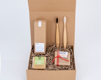 May include: A brown cardboard box with a lid open, revealing a gift set. The set includes a bottle of peppermint tea, a bar of soap, a small box of orange-flavored candy, and two bamboo toothbrushes.