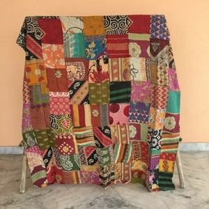 Könnte beinhalten: Ein Patchwork-Quilt mit einer Vielzahl von bunten Stoffquadraten. Der Quilt weist eine Mischung aus Mustern auf, darunter Blumen-, geometrische und abstrakte Designs. Die Farbpalette umfasst Rot-, Orange-, Grün-, Blau- und Lilatöne. Der Quilt ist über einem Holzrahmen drapiert.
