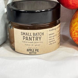 May include: A small glass jar with a black lid, labeled "SMALL BATCH PANTRY" and "Spiced APPLE PIE BUTTER." The jar contains a brown, spreadable substance. Two red and yellow apples are in the background.