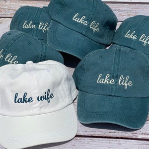 K&ouml;nnte beinhalten: F&uuml;nf blaugr&uuml;ne Baseballkappen mit dem gestickten Schriftzug "lake life" in Wei&szlig;. Eine wei&szlig;e Baseballkappe mit dem gestickten Schriftzug "lake wife" in Blau.