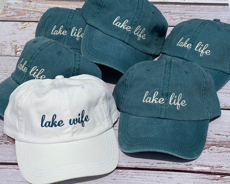 K&ouml;nnte beinhalten: F&uuml;nf blaugr&uuml;ne Baseballkappen mit dem gestickten Schriftzug "lake life" in Wei&szlig;. Eine wei&szlig;e Baseballkappe mit dem gestickten Schriftzug "lake wife" in Blau.