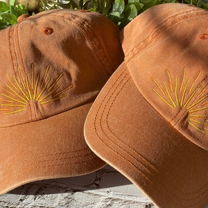 May include: Two brown baseball caps with yellow embroidered sun designs. The caps are made of a soft, textured fabric.