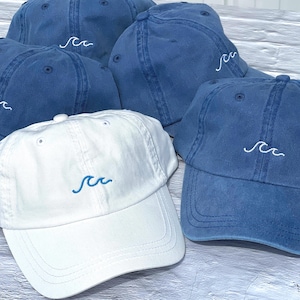 May include: A collection of baseball caps in blue and white. The blue caps feature white embroidered wave designs, while the white cap features a blue embroidered wave design. Each cap has a curved brim, a button top, and stitched eyelets.