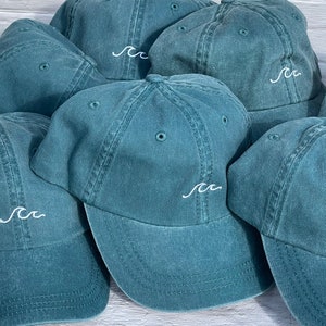 May include: A group of teal blue baseball caps with a white embroidered wave design. The caps are arranged in a semi-circle on a white wooden surface.