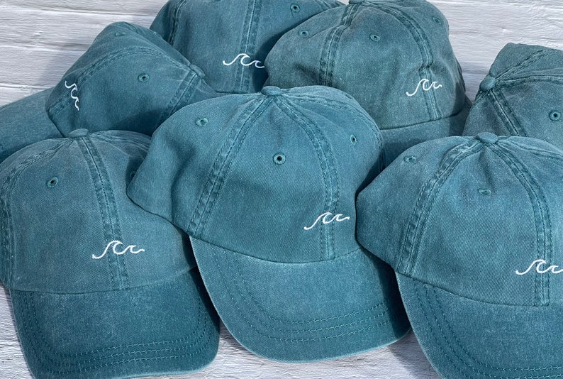 May include: A group of teal blue baseball caps with a white embroidered wave design. The caps are arranged in a semi-circle on a white wooden surface.
