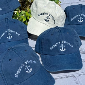 May include: A collection of navy blue and one off-white baseball caps. Each cap features the embroidered text "Maggie's Vineyard" above a white anchor. The caps are made of a soft, washed cotton material.