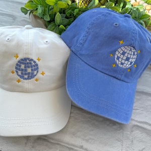 May include: Two baseball caps with a disco ball design embroidered on the front. One cap is white and the other is blue.