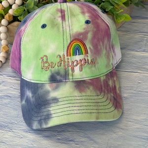 May include: A green, purple, and blue tie-dye baseball cap with an embroidered rainbow and the text "Be a Hippie".
