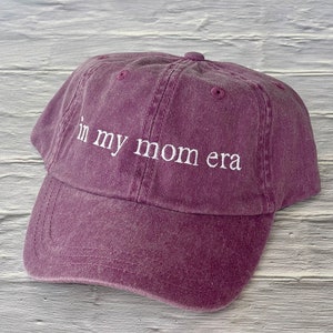 May include: A purple baseball cap with the text "in my mom era" embroidered in white.