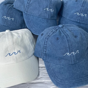 May include: Five denim and white baseball caps with a wave design embroidered on the front. The caps are arranged in a pile on a wooden surface.