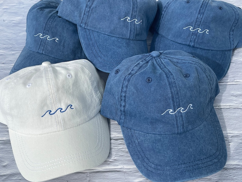 May include: Five denim and white baseball caps with a wave design embroidered on the front. The caps are arranged in a pile on a wooden surface.