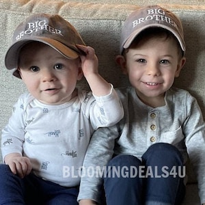 May include: Two young boys wearing gray baseball caps with the text "Big Brother" embroidered on them. The boys are sitting on a couch and smiling at the camera.