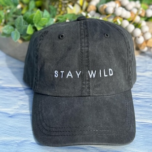 以下が含まれることがあります： 白い刺繍で「STAY WILD」と書かれた黒いコットン製のベースボールキャップです。