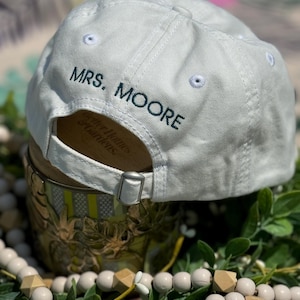 May include: A light gray baseball cap with the text "MRS. MOORE" embroidered in dark green. The cap has a curved brim, a metal clasp, and white stitching details. The cap is a classic style, suitable for everyday wear.
