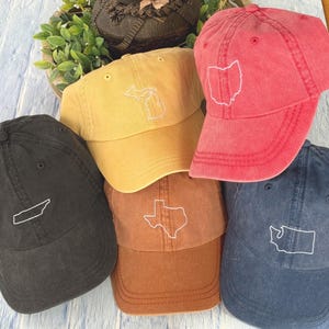 May include: Five different colored baseball caps with embroidered state outlines. The caps are black, red, yellow, brown, and blue. The states are Tennessee, Ohio, Michigan, Texas, and Washington.