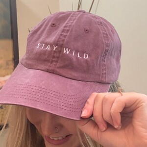 以下が含まれることがあります： 前面に白い刺繍で「STAY WILD」と書かれた紫色のベースボールキャップ