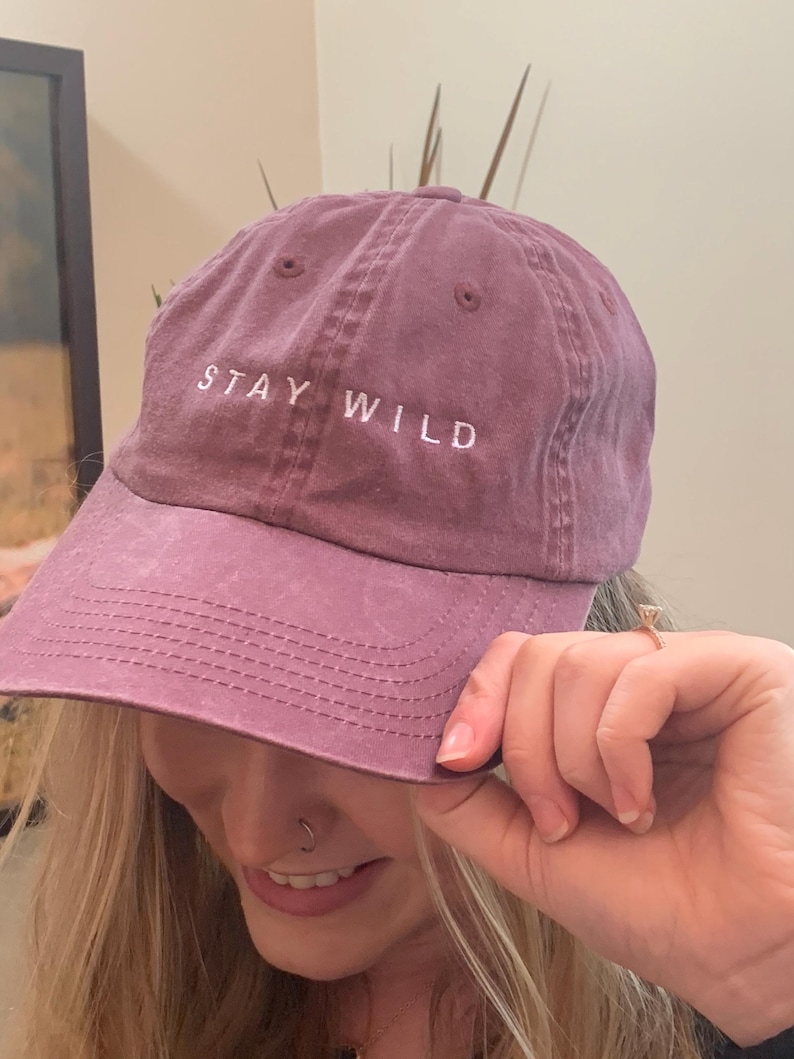 以下が含まれることがあります： 前面に白い刺繍で「STAY WILD」と書かれた紫色のベースボールキャップ