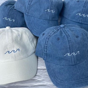 May include: A collection of baseball caps in various shades of blue and one in off-white. Each cap features a simple embroidered wave design on the front panel. The blue caps have white wave stitching, while the off-white cap has blue wave stitching.