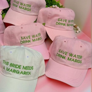 May include: Five pink baseball caps with green embroidery that reads "Save Water Drink Margs" and one white baseball cap with green embroidery that reads "This Bride Needs a Margarita".