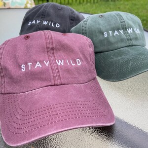 以下が含まれることがあります： 前面に「Stay Wild」という刺繍が施された野球帽が3つあります。帽子は、ワインレッド、ダークグリーン、ブラックです。