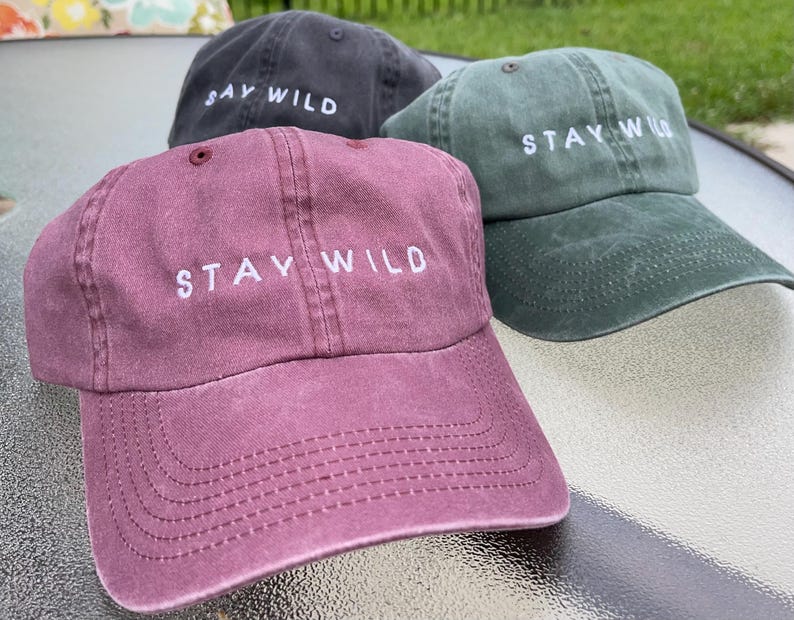 以下が含まれることがあります： 前面に「Stay Wild」という刺繍が施された野球帽が3つあります。帽子は、ワインレッド、ダークグリーン、ブラックです。