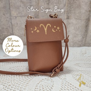 May include: A brown faux leather crossbody bag with a gold chain strap and a gold zipper. The front flap of the bag has a gold Aries star sign with moons and stars.