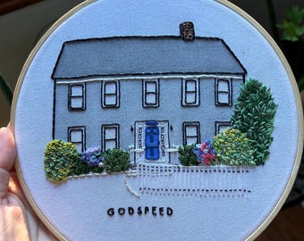 May include: Embroidered hoop art depicting a grey house with a blue door and white picket fence. The house has four windows and a chimney. The hoop is surrounded by green and yellow foliage. The text "GODSPEED" is embroidered below the house.