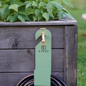 May include: A green metal hose holder with a brass faucet is mounted on a wooden planter box. The holder has the word "LIVIO" printed on it. A black hose with orange accents is coiled below the holder, and green plants are growing in the planter.