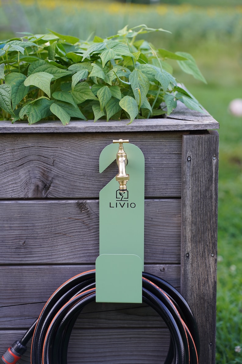 May include: A green metal hose holder with a brass faucet is mounted on a wooden planter box. The holder has the word "LIVIO" printed on it. A black hose with orange accents is coiled below the holder, and green plants are growing in the planter.