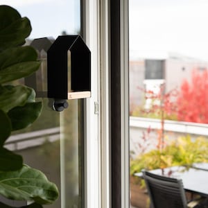 Op de afbeelding: Een zwarte vogelvoederbak in de vorm van een huisje met een lichte houten bodem, bevestigd aan een raam met een ronde zuignap. De voederbak heeft een kleine zwarte knop aan de onderkant en een logo op de houten rand aan de voorzijde.