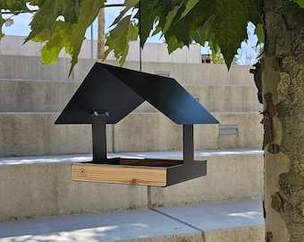May include: Black metal bird feeder with a wooden tray and a roof. The feeder is hanging from a tree branch and says "Livio" on the tray.