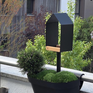 May include: A black metal bird feeder in a house shape, with a wooden perch, mounted on a black pole. It is set against a backdrop of greenery and a building. A black planter with green plants is in the foreground.