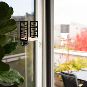 Puede incluir: Una estantería de pared de metal negro y madera con un diseño geométrico. La estantería está fijada a un marco de ventana y tiene una pequeña base de madera de color claro. El fondo muestra un balcón con plantas y una mesa.