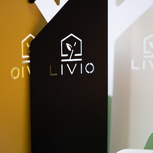May include: Close-up of three rectangular signs with the word "LIVIO" in a modern font. The signs are in black, gold, and off-white, with a logo of a house and a plant. The background is blurred, with hints of green and white.