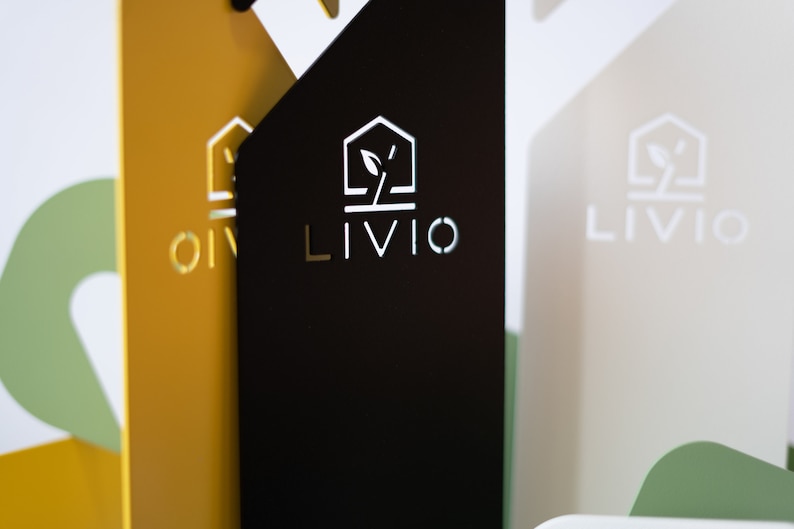May include: Close-up of three rectangular signs with the word "LIVIO" in a modern font. The signs are in black, gold, and off-white, with a logo of a house and a plant. The background is blurred, with hints of green and white.