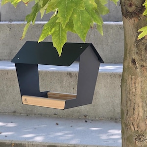 以下が含まれることがあります： マットブラックの金属製バードフィーダー。傾斜のある屋根と明るい色の木製ベースが特徴です。木製プラットフォームの前面には、小さな家の形をしたロゴアイコンと共に、ブランド名「LIVIO」が黒い文字でプリントされています。