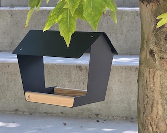 May include: A matte black metal bird feeder with a pitched roof design and a light-colored wooden base. The front edge of the wooden platform features the brand name LIVIO printed in black text alongside a small house-shaped logo icon.