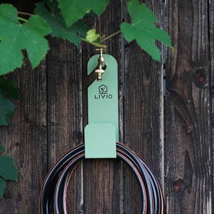 May include: A mint green hose holder with a brass faucet is mounted on a weathered wooden fence. The holder has the word "LIVIO" printed on it. A black and orange hose is coiled below, with green leaves in the background.