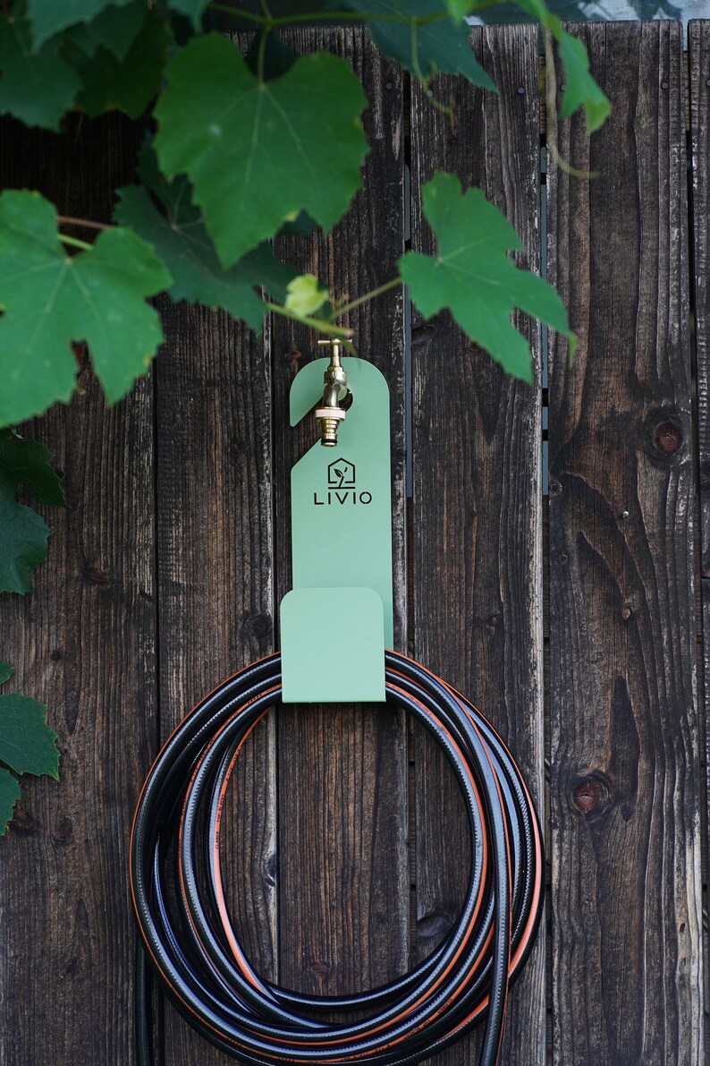 May include: A mint green hose holder with a brass faucet is mounted on a weathered wooden fence. The holder has the word "LIVIO" printed on it. A black and orange hose is coiled below, with green leaves in the background.