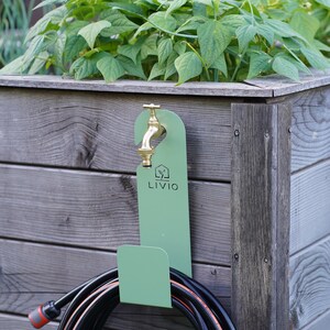 May include: A green metal hose holder with a brass faucet and a coiled black hose. The holder is mounted on a wooden planter box filled with green plants. The text "LIVIO" is visible on the holder.