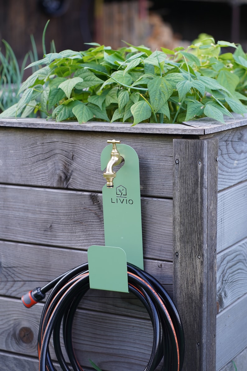 May include: A green metal hose holder with a brass faucet and a coiled black hose. The holder is mounted on a wooden planter box filled with green plants. The text "LIVIO" is visible on the holder.